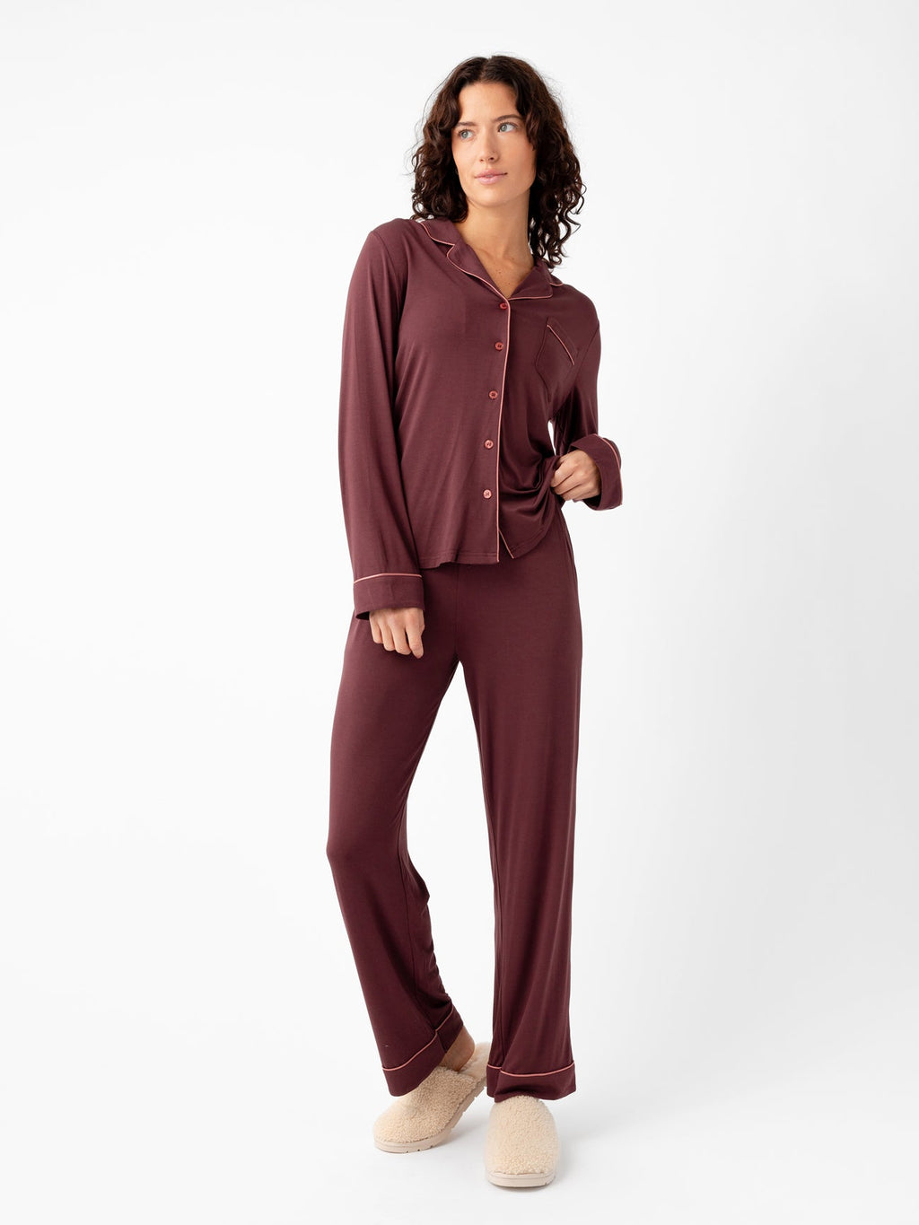 A person with curly hair stands against a white background, wearing Cozy Earth's Last Chance Women's Bamboo Stretch-Knit Long Sleeve Pajama Set in maroon with pink piping and holds the shirt's hem. They're also wearing beige slippers. |Color:Burgundy