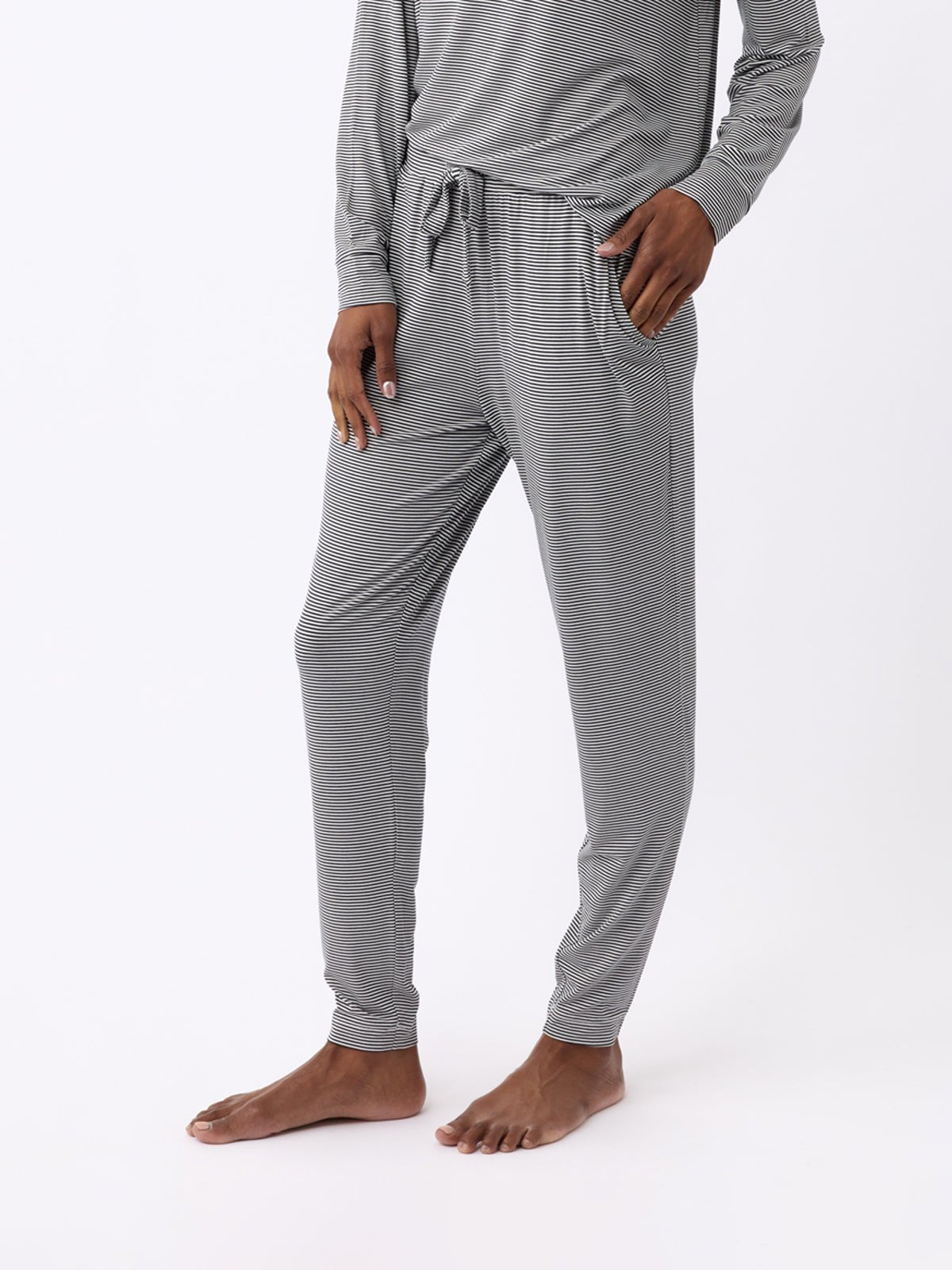 A person models Cozy Earth's Women's Bamboo Stretch-Knit Tapered Pants, featuring gray stripes and a matching long-sleeved top. The photo highlights the pants' lower half against a plain white background, with one hand in a pocket. |Color:Eclipse Mini Stripe