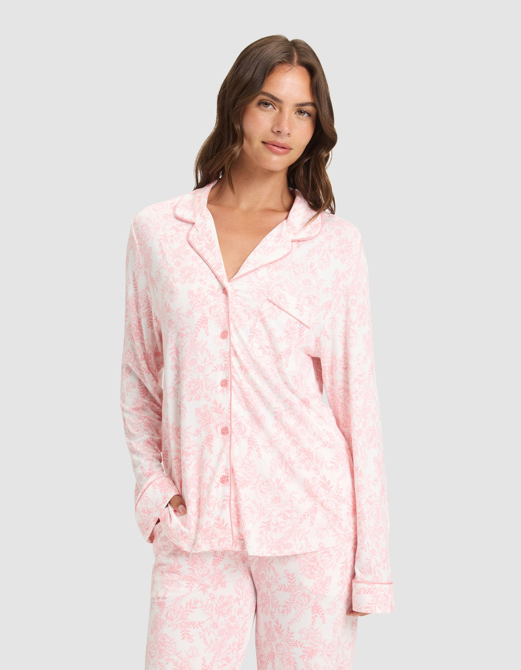 A woman models the Cozy Earth Women's Bamboo Stretch-Knit Long Sleeve Pajama Set in light pink and white, standing against a plain light gray background with one hand in her pocket, smiling softly at the camera. |Color:Tulip Toile