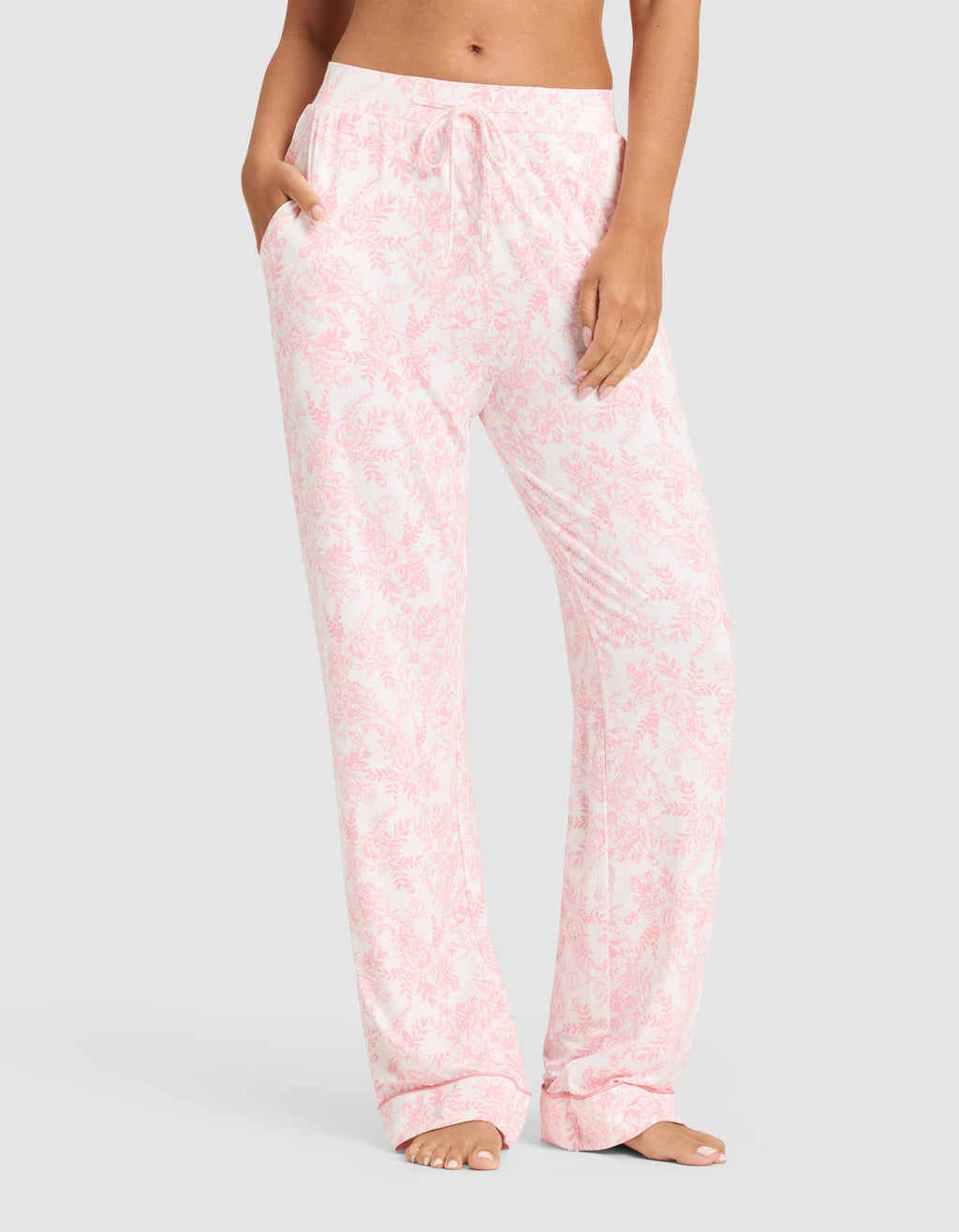 A woman wears Cozy Earth’s Women's Bamboo Stretch-Knit Long Sleeve Pajama Set in light pink and white floral, featuring relaxed-fit pants with pockets and a drawstring waist, standing barefoot against a plain background. |Color:Tulip Toile