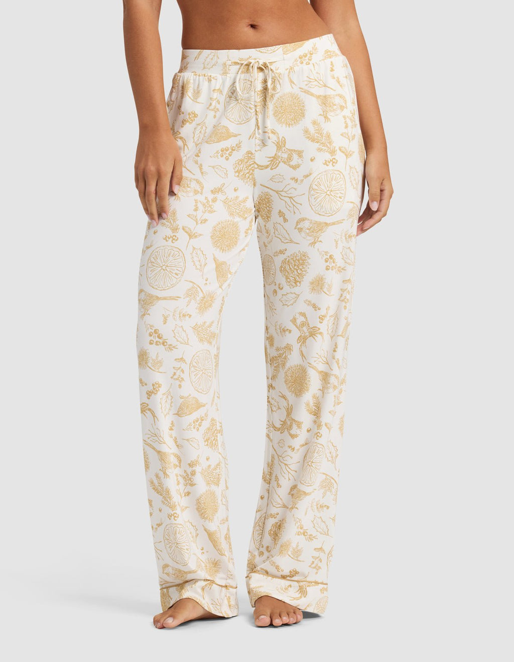 A person stands barefoot against a plain light background wearing Cozy Earth's Women's Bamboo Stretch-Knit Pajama Pant in a white color with a yellow floral and fruit pattern.|Color:Winter Toile Dijon