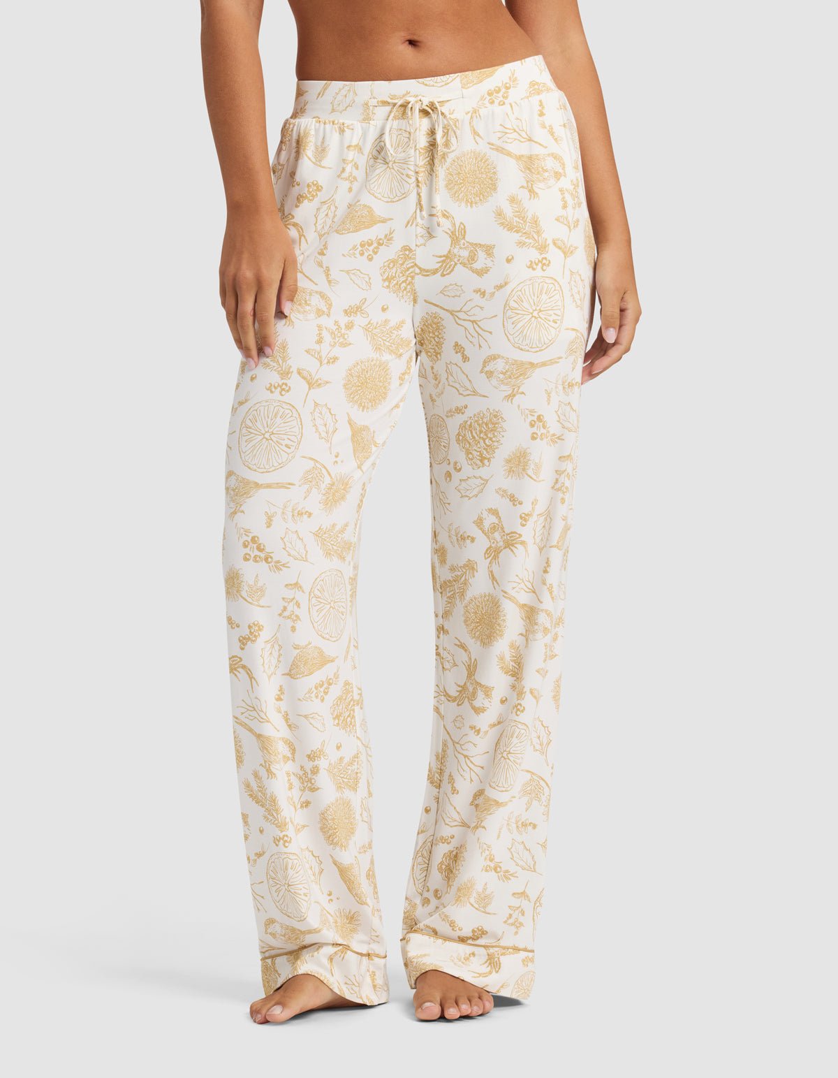 A person stands barefoot against a plain light background wearing Cozy Earth's Women's Bamboo Stretch-Knit Pajama Pant in a white color with a yellow floral and fruit pattern.|Color:Winter Toile Dijon