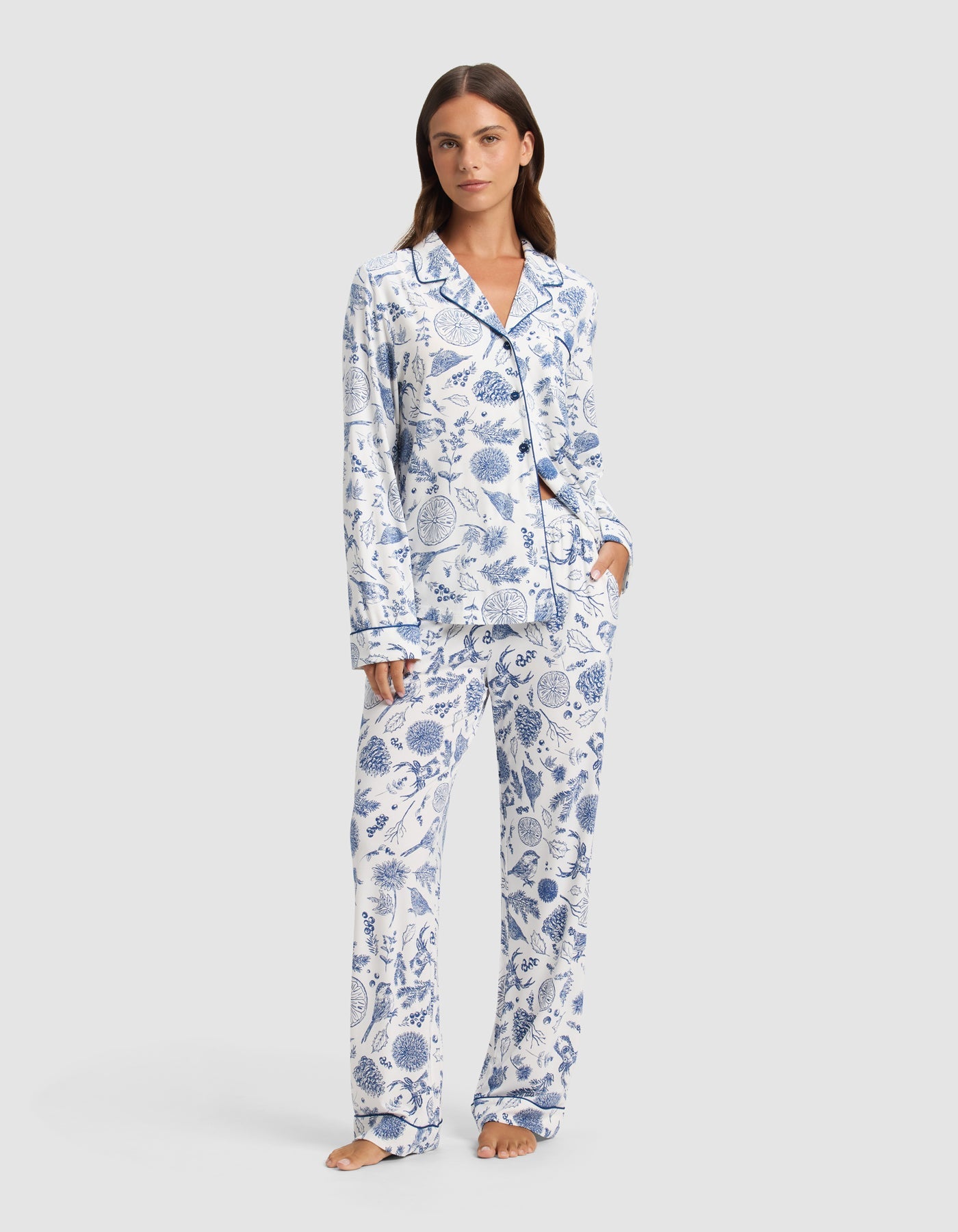 A woman stands barefoot wearing Cozy Earth's Women's Bamboo Stretch-Knit Pajama Pant, featuring a white background with blue botanical print and paired with a matching long-sleeve button-up top.|Color:Winter Toile Pacific