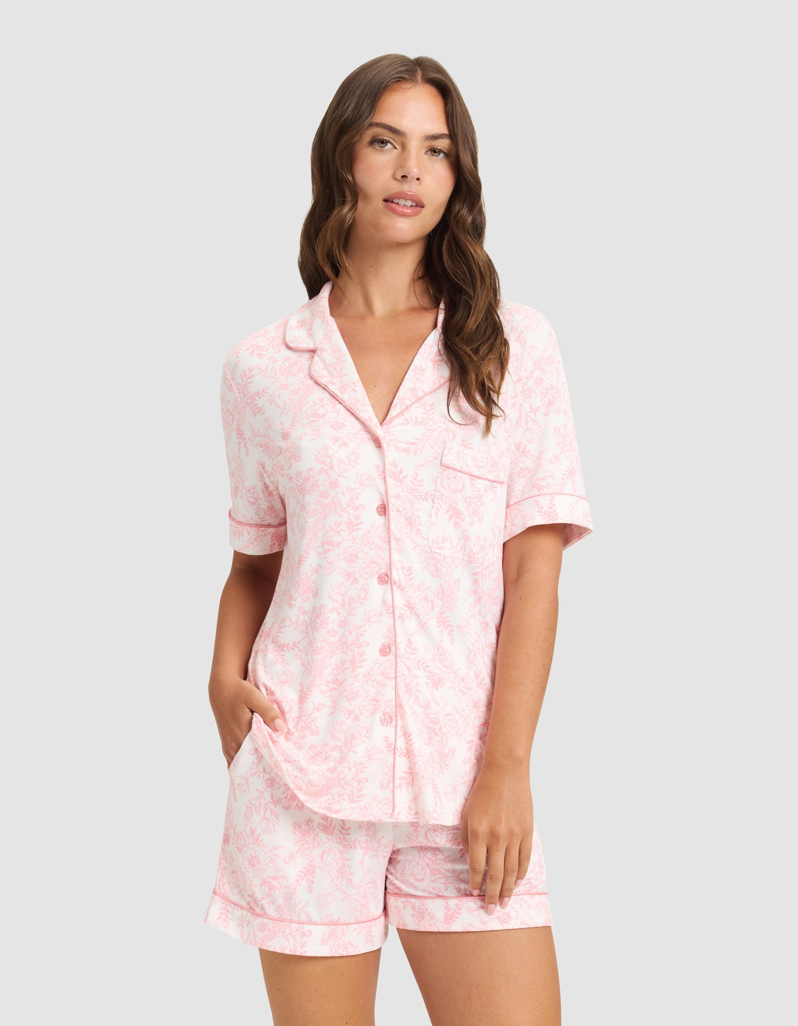 A woman with long brown hair models the HIDE Women's Bamboo Stretch-Knit Short Sleeve Pajama Top in light pink, featuring a subtle floral pattern, button-up front, and matching shorts, standing against a plain light background. |Color:Tulip Toile