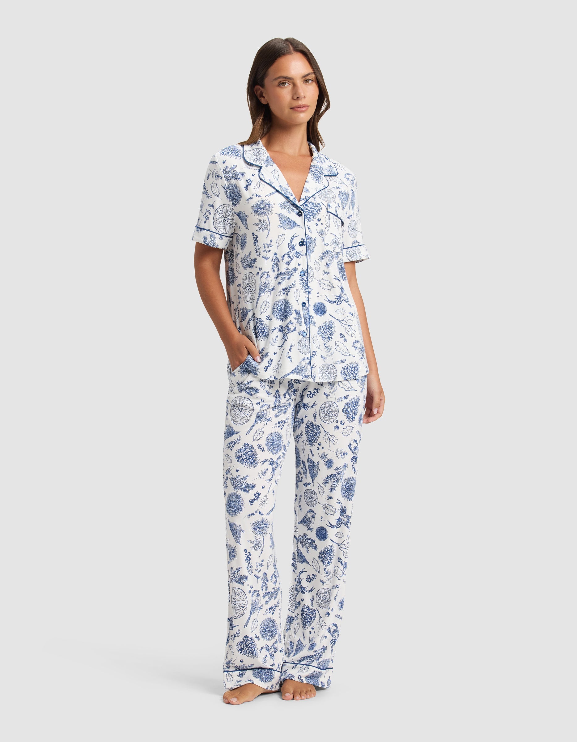 A woman stands barefoot in the Cozy Earth Women's Bamboo Stretch-Knit Short Sleeve Pajama Set, featuring a blue and white botanical print with a collared button-up top and loose pants. She has one hand in her pocket and looks relaxed.|Color:Winter Toile Pacific