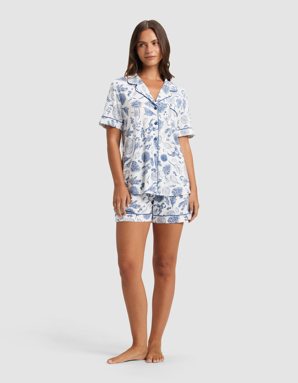 A woman poses barefoot against a light gray backdrop, wearing the Cozy Earth Women's Bamboo Stretch-Knit Short Sleeve Pajama Set—a matching button-up top with a collar and shorts, featuring a blue botanical print on white.|Color:Winter Toile Pacific
