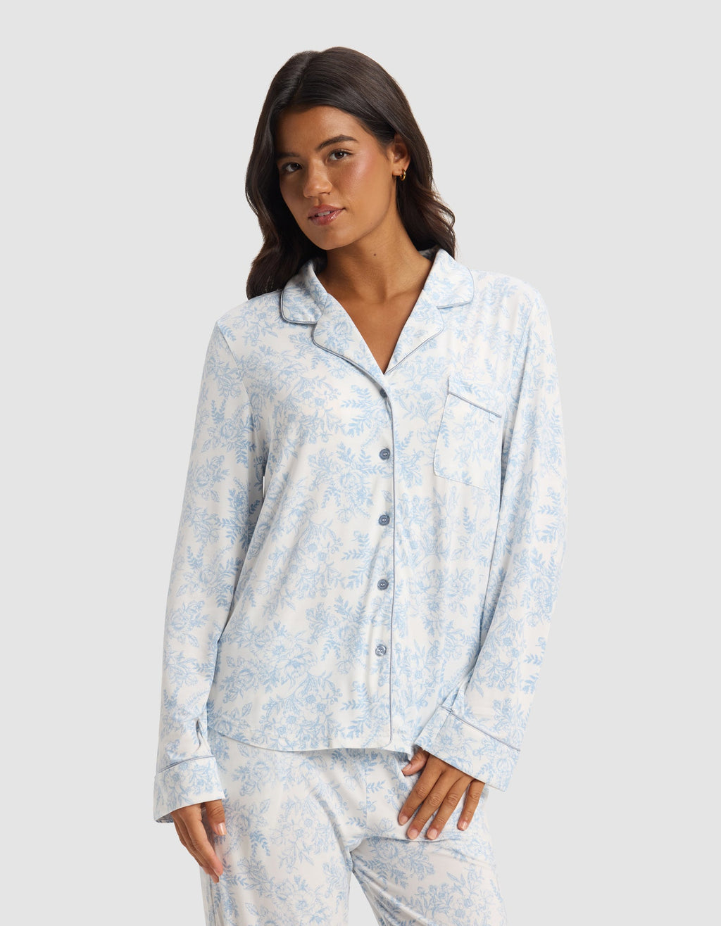 A woman wears the Cozy Earth Women's Bamboo Stretch-Knit Long Sleeve Pajama Set in light blue and white print, featuring a button-up top and matching pants, standing against a plain light gray background.|Color:Sky Toile