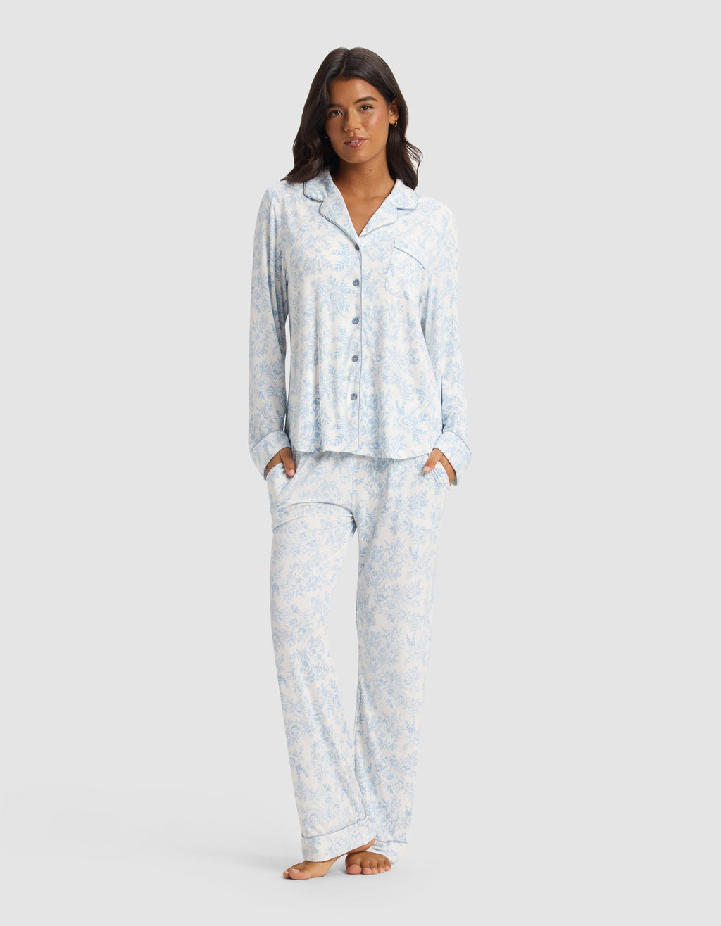 A woman stands barefoot, hands in her pockets, wearing the Cozy Earth Women's Bamboo Stretch-Knit Long Sleeve Pajama Set in a light blue and white pattern, against a plain light gray background.|Color:Sky Toile