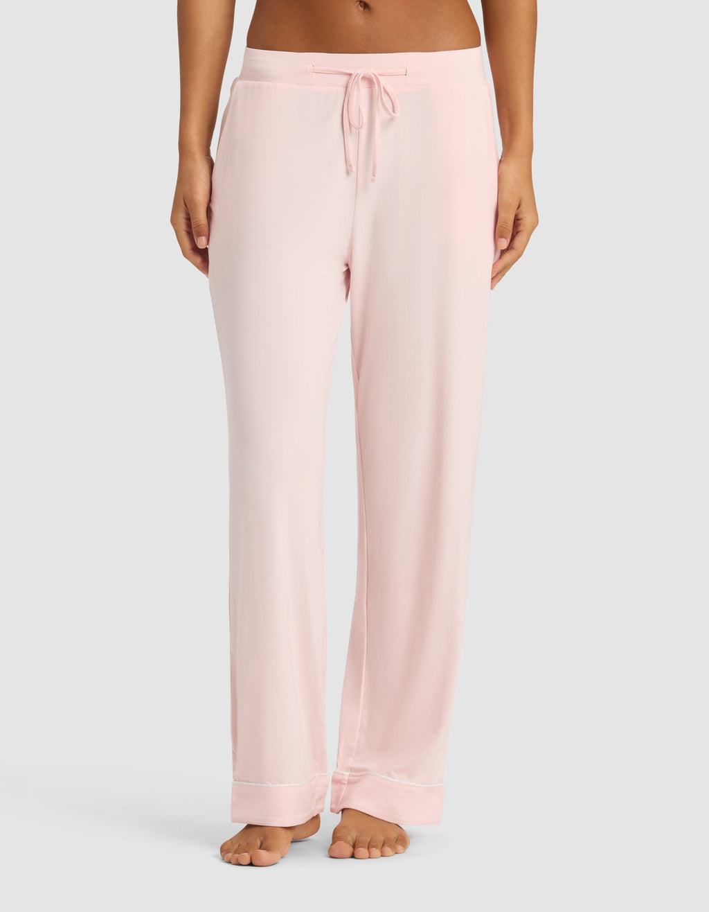 A woman wears Cozy Earth's Women's Bamboo Stretch-Knit Long Sleeve Pajama Set in light pink with a drawstring waist, standing barefoot against a plain light gray background.|Color:Blossom