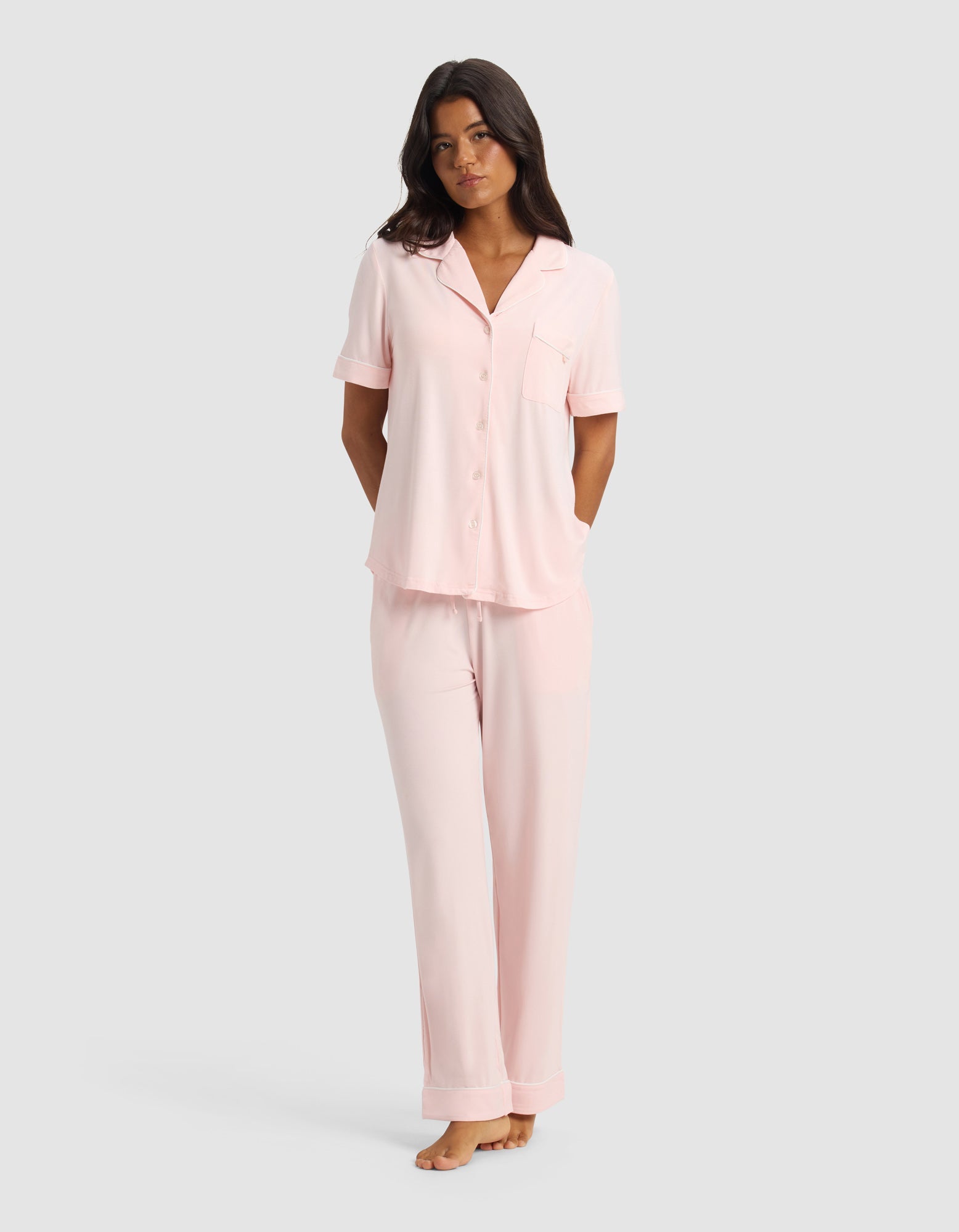 A woman stands barefoot against a plain light gray background, wearing the Cozy Earth Women's Bamboo Stretch-Knit Short Sleeve & Pant Pajama Set in Blossom, with her arms relaxed behind her back.|Color:Blossom