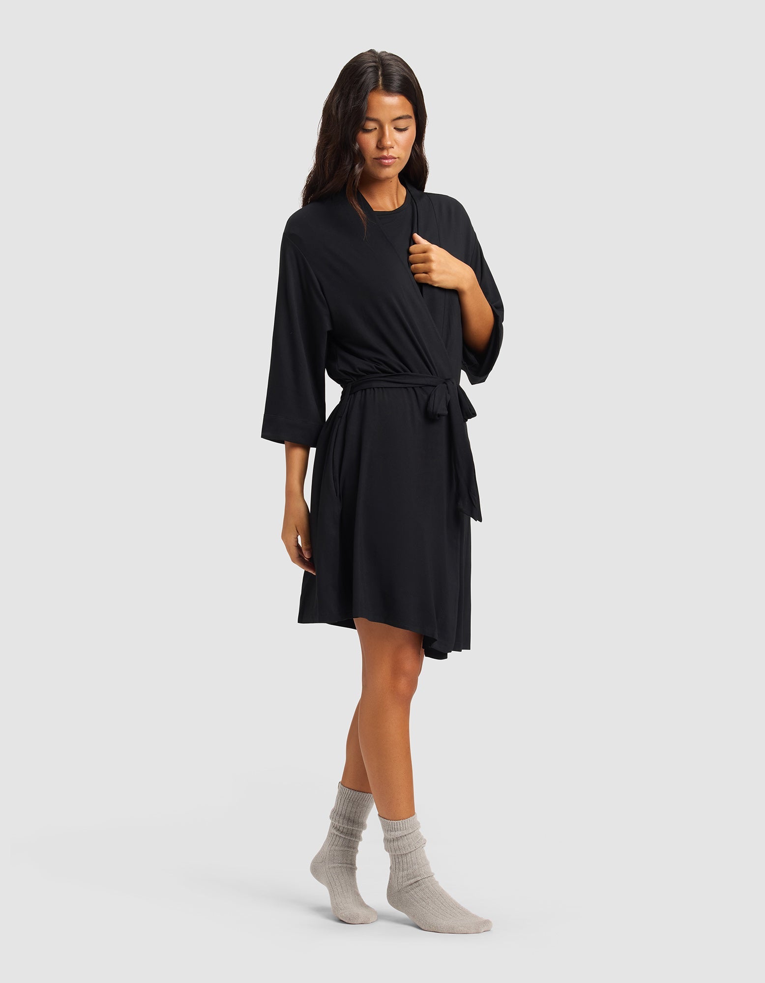 A woman with long dark hair stands against a plain background wearing the Cozy Earth Women's Bamboo Stretch-Knit Kimono Robe in black, featuring three-quarter sleeves and a tie waist, paired with light gray socks. She has a relaxed posture.|Color:Black