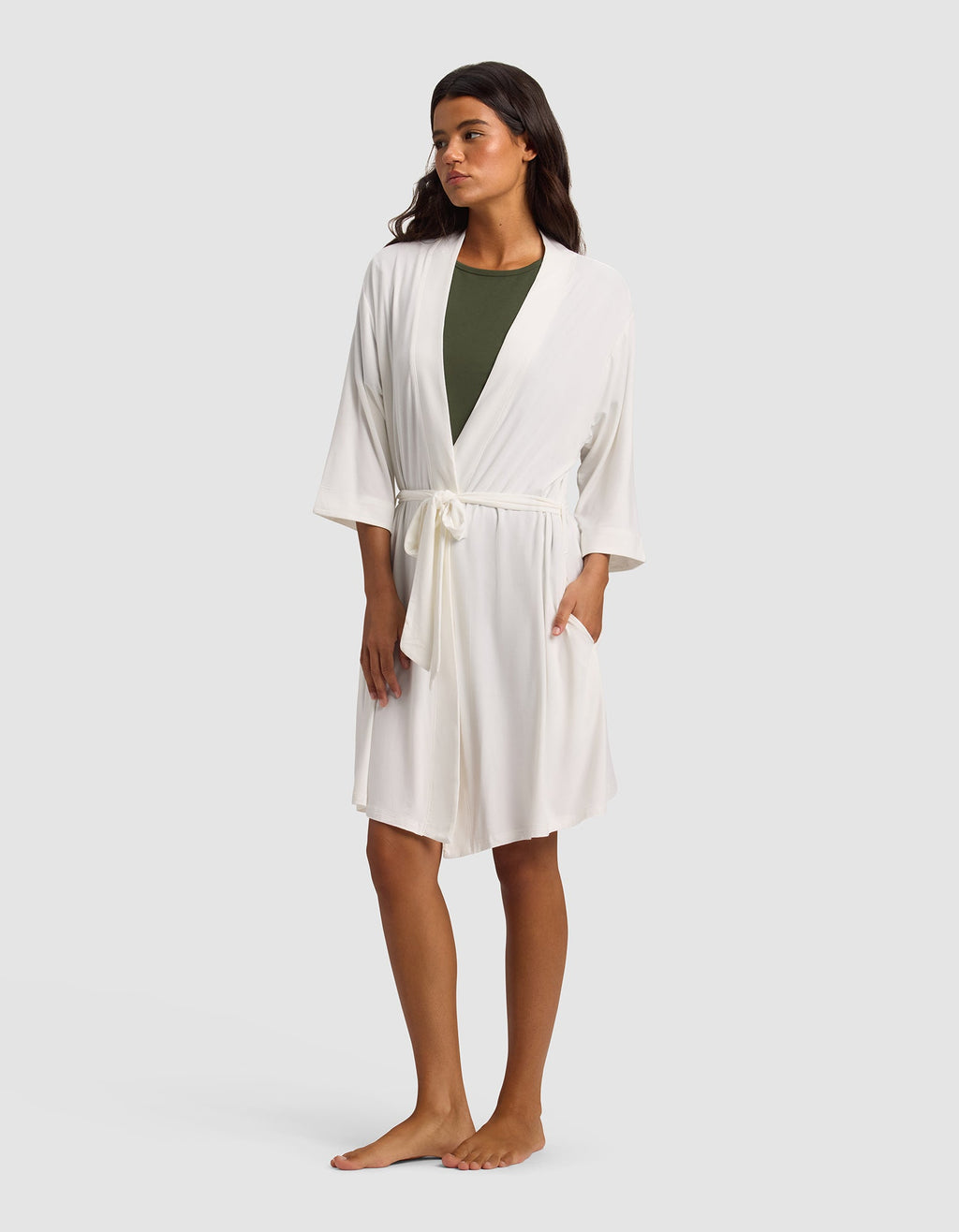 A woman stands barefoot against a light gray background, wearing Cozy Earth's Women's Bamboo Stretch-Knit Kimono Robe in white with a waist tie over an olive green top.|Color:Ivory