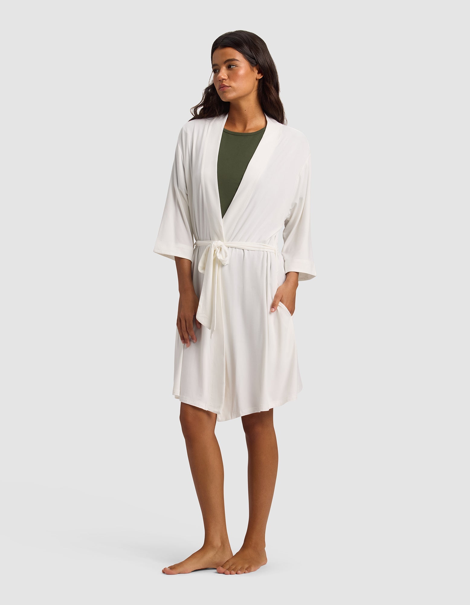 A woman stands barefoot against a light gray background, wearing Cozy Earth's Women's Bamboo Stretch-Knit Kimono Robe in white with a waist tie over an olive green top.|Color:Ivory