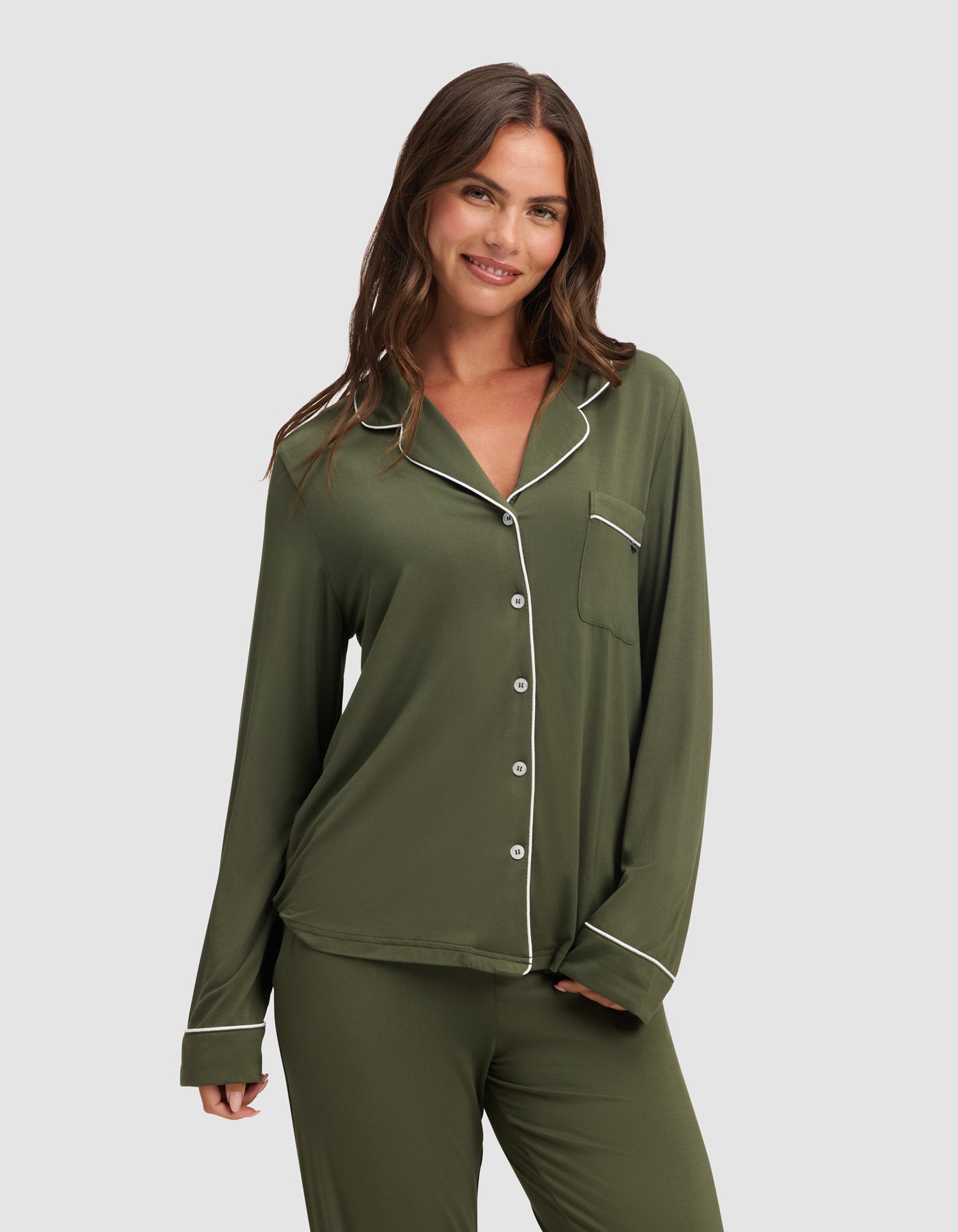 A woman with long brown hair smiles while wearing the HIDE Women's Bamboo Stretch-Knit Long Sleeve Pajama Top in olive green with white piping, paired with matching pants, standing against a plain light gray background. |Color:Olive