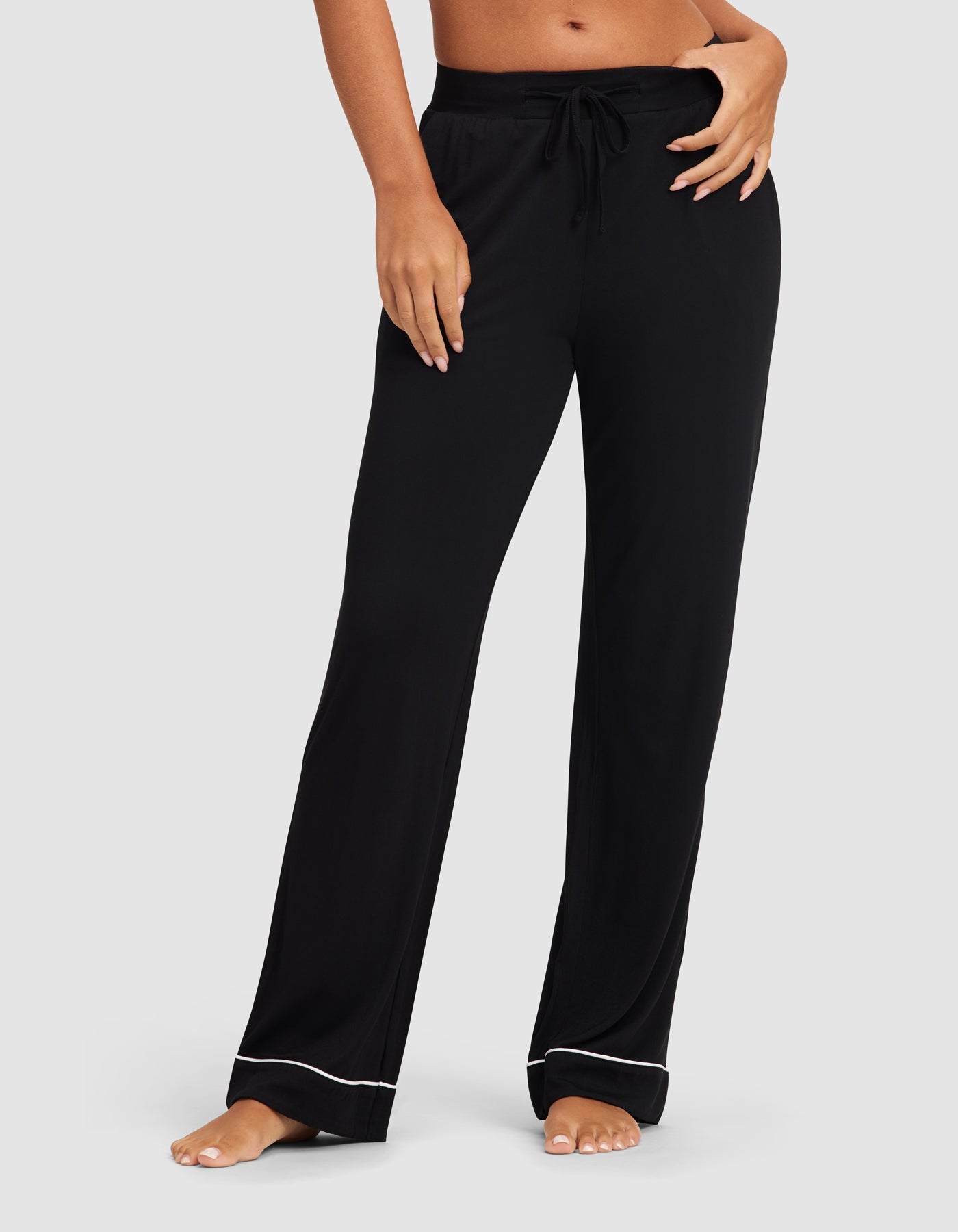 A woman models the Cozy Earth Women's Bamboo Stretch-Knit Pajama Pant in black, featuring a drawstring waist and white piping at the hem, standing barefoot against a light gray background.|Color:Black