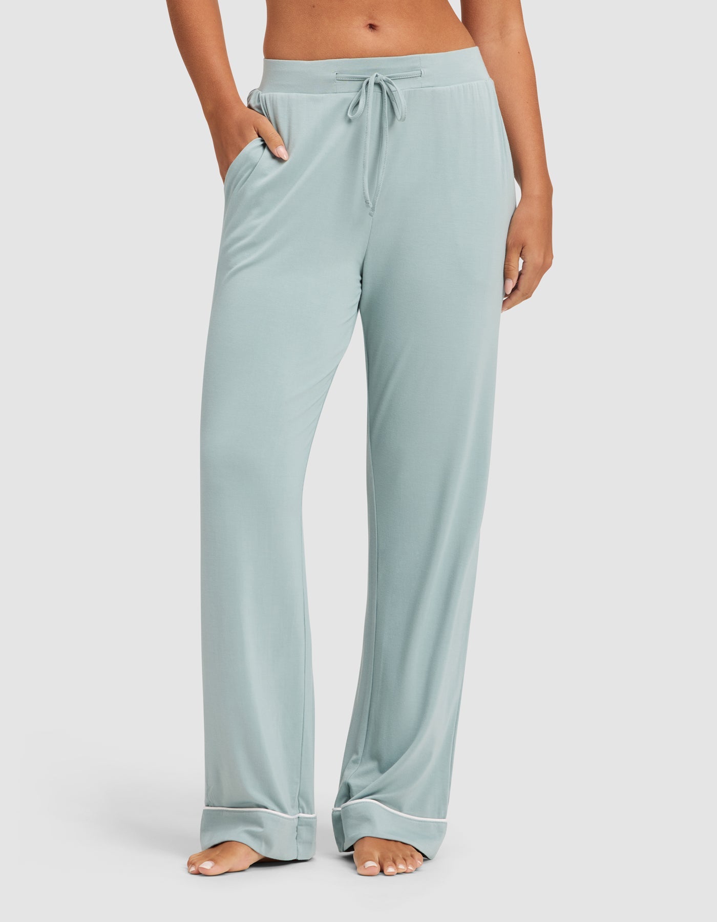 A person stands barefoot with hands in the pockets of Cozy Earth Women's Bamboo Stretch-Knit Pajama Pant in light blue. The relaxed-fit, drawstring pants have side pockets, and the photo is cropped at their waist and ankles. |Color:Brook