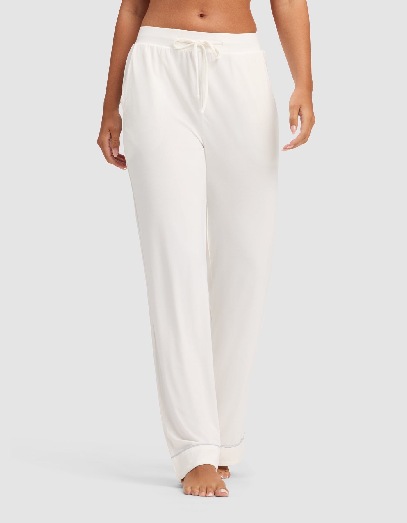 A model wears HIDE Women's Bamboo Stretch-Knit Pajama Pants in white, featuring a loose fit and drawstring waist, barefoot against a plain light gray background. Only the lower torso and legs are shown.|Color:Ivory