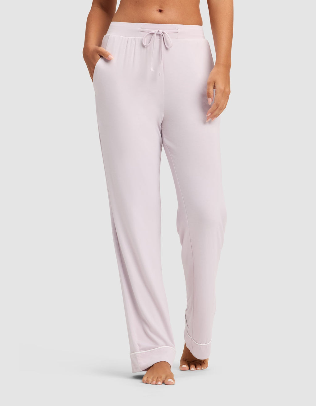 A woman wears HIDE's Women's Bamboo Stretch-Knit Pajama Pant in light pink, featuring a drawstring waist and side pockets, standing barefoot against a plain background. |Color:Lilac