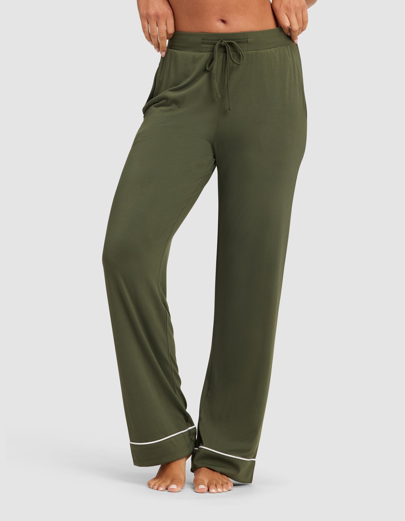 A woman models the HIDE Women's Bamboo Stretch-Knit Pajama Pant in olive green, featuring a drawstring waist and white hem piping, while standing barefoot against a plain white background. |Color:Olive