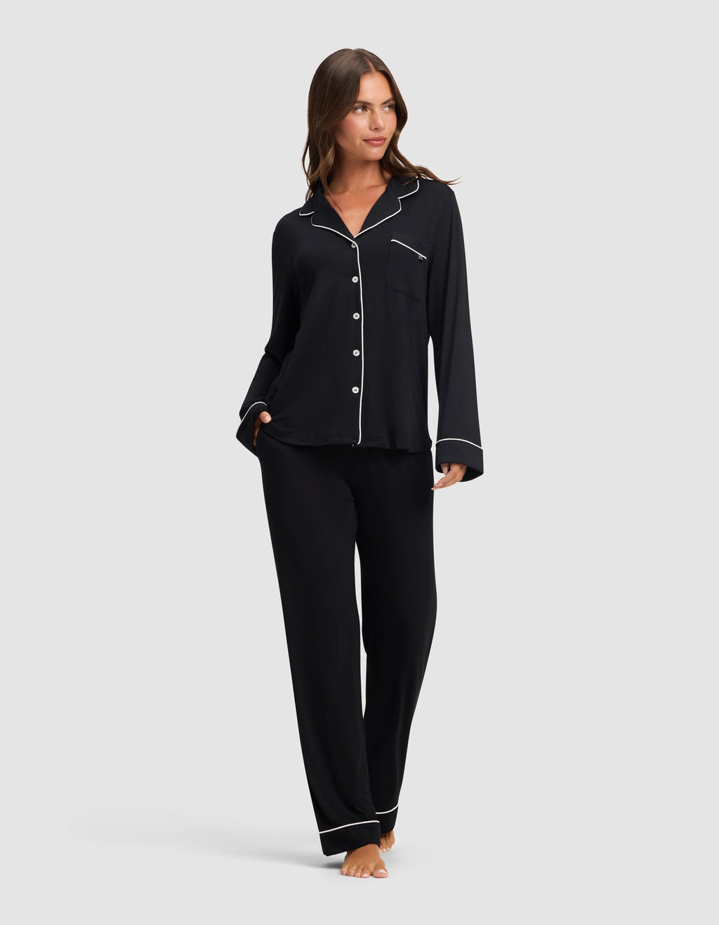 A woman stands barefoot against a light gray background, wearing Cozy Earth's Women's Bamboo Stretch-Knit Long Sleeve Pajama Set in black with white piping, featuring a button-up top and matching pants. |Color:Black