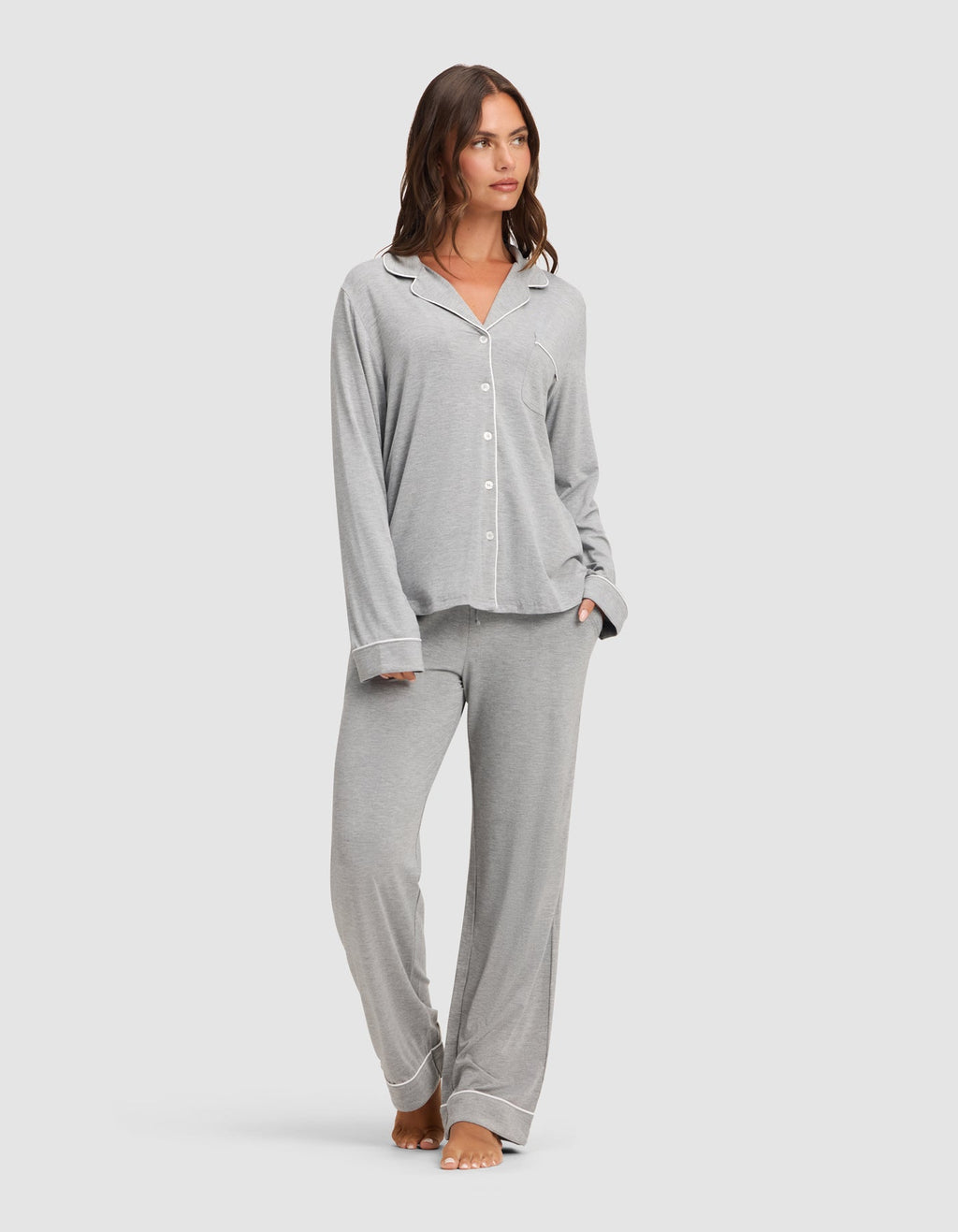 A woman stands barefoot against a plain light background, wearing the Cozy Earth Women's Bamboo Stretch-Knit Long Sleeve Pajama Set in light grey with white piping, featuring a button-up top and matching pants.  |Color:Grey
