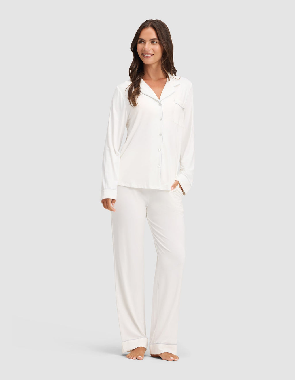 A woman smiles while wearing the Cozy Earth Women's Bamboo Stretch-Knit Long Sleeve Pajama Set, featuring a button-up top. She stands barefoot against a plain light gray background. |Color:Ivory
