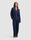 A woman poses barefoot against a plain light background, smiling in the Cozy Earth Women's Bamboo Stretch-Knit Long Sleeve Pajama Set—navy blue with white piping, featuring a button-up top and loose-fitting pants. |Color:Navy