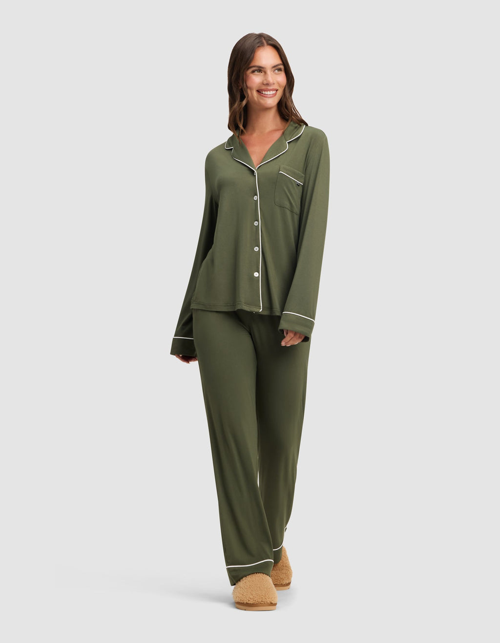 A woman smiles against a light gray background, wearing Cozy Earth's Women's Bamboo Stretch-Knit Long Sleeve Pajama Set in olive green with white piping and beige slippers.|Color:Olive
