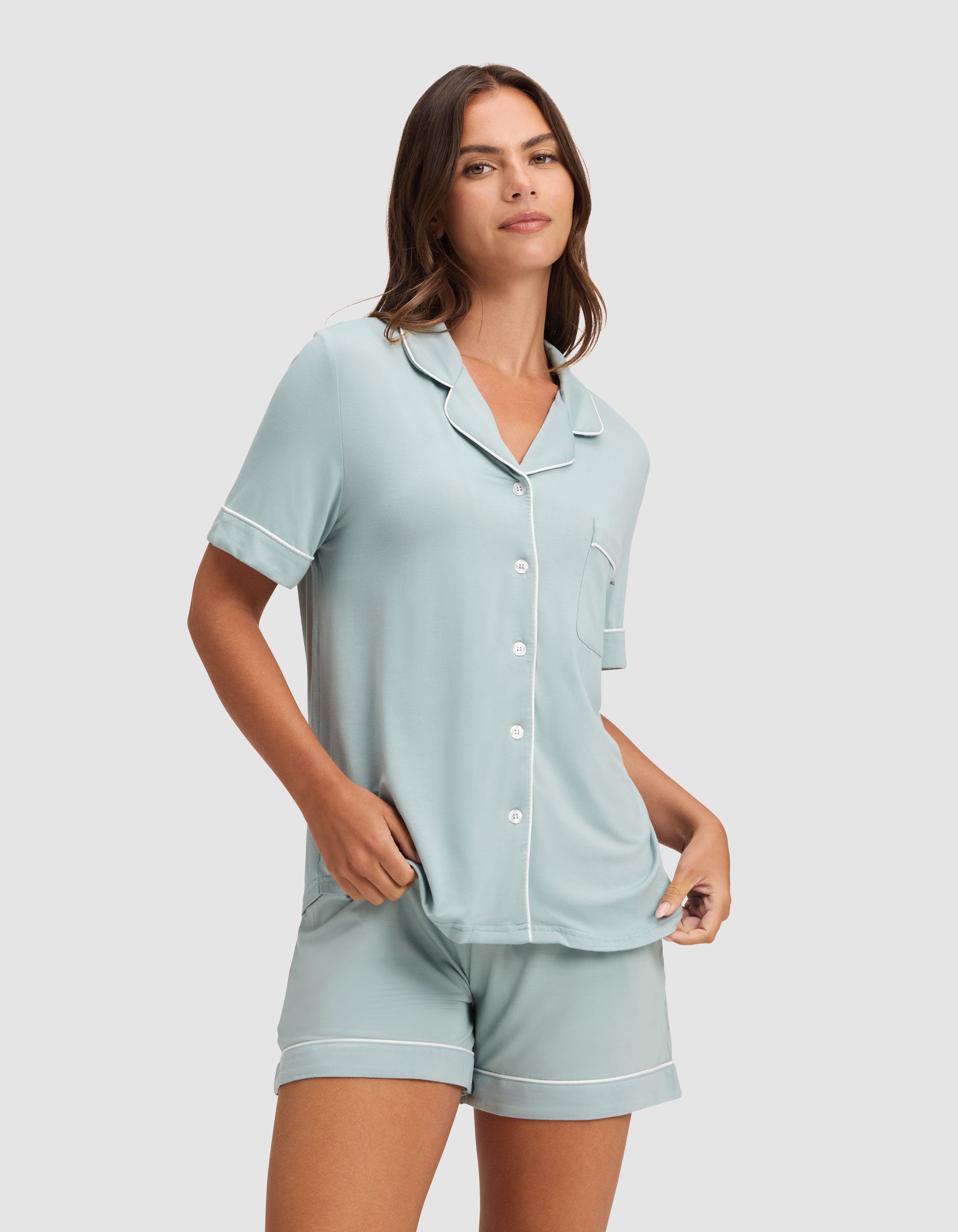 A woman stands against a plain light gray background wearing the Cozy Earth Women's Bamboo Stretch-Knit Short Sleeve Pajama Set in light blue with white trim, featuring a button-up top and matching shorts. |Color:Brook