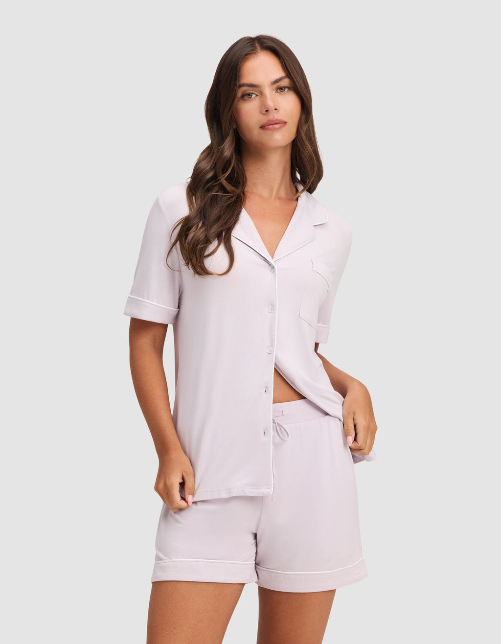 A woman with long brown hair wears the HIDE Women's Bamboo Stretch-Knit Pajama Short set with a button-up top and matching shorts in light lavender, standing against a neutral background.|Color:Lilac