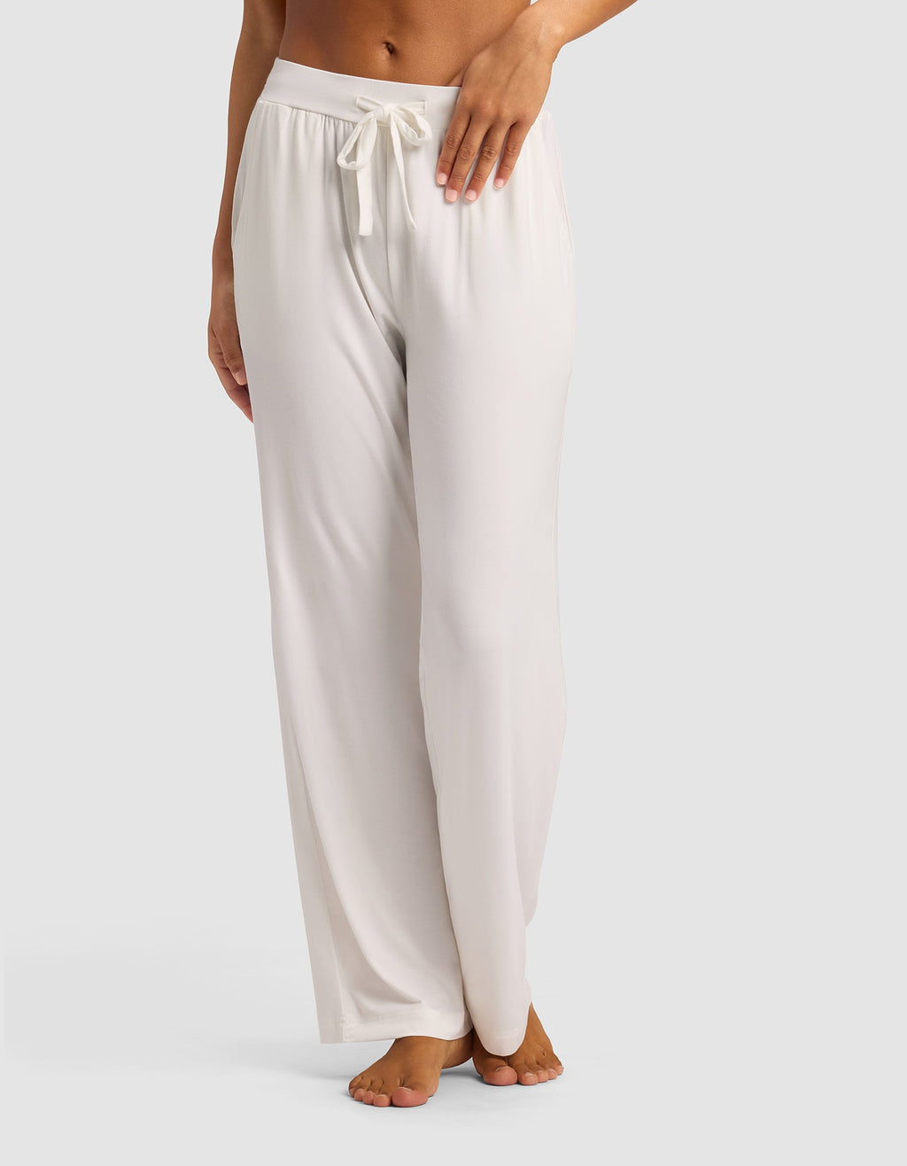 A woman stands barefoot against a plain light background, modeling the lower half of Cozy Earth's Women's Bamboo Stretch-Knit Pant—loose-fitting, light-colored lounge pants with a drawstring waist.|Color:Ivory