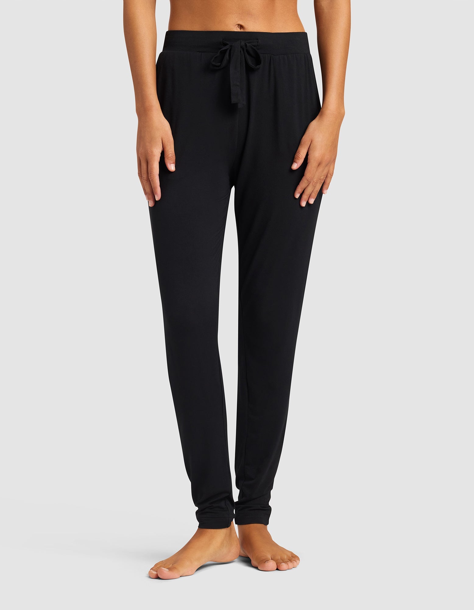 A woman stands barefoot against a light gray background, wearing Cozy Earth's Women's Bamboo Stretch-Knit Tapered Pant in black. Her hands rest at her sides, and the focus is on the pants from waist to ankles.|Color:Black
