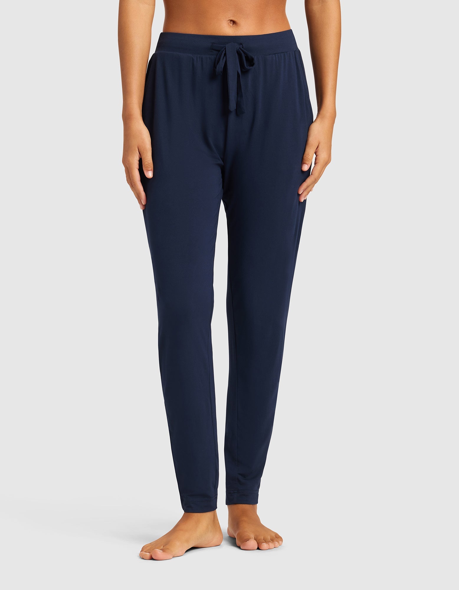 A woman models Cozy Earth's Women's Bamboo Stretch-Knit Tapered Pant in navy, shown barefoot with only her lower body visible against a plain light grey background.|Color:Navy