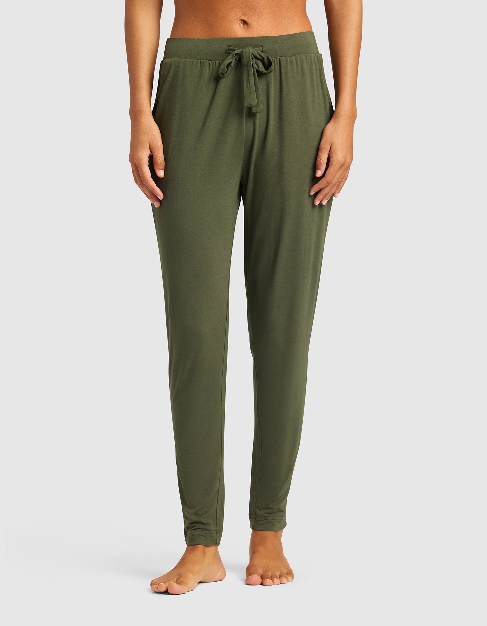 A person stands barefoot against a plain white background, wearing Cozy Earth's Women's Bamboo Stretch-Knit Tapered Pant in olive green with a drawstring waist and bare midriff, shown from shoulders to feet.|Color:Olive