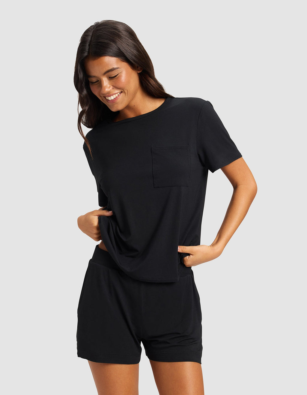 A woman with long dark hair wears the Cozy Earth Women's Bamboo Stretch-Knit Lounge Tee in black, paired with matching shorts. She smiles and looks down, hands in her pockets, standing against a light gray background.|Color:Black