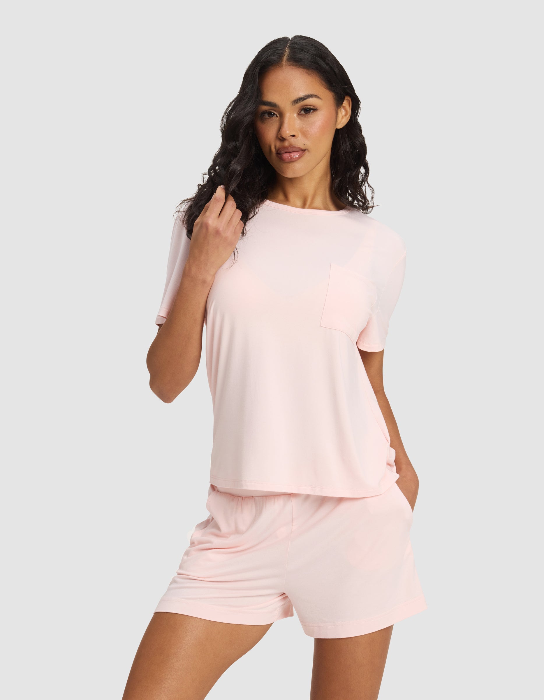 A woman with long, wavy dark hair and a relaxed, confident look stands against a plain background wearing Cozy Earth's Women's Bamboo Stretch-Knit Lounge Tee in light pink with a pocket, paired with matching pink shorts.|Color:Blossom